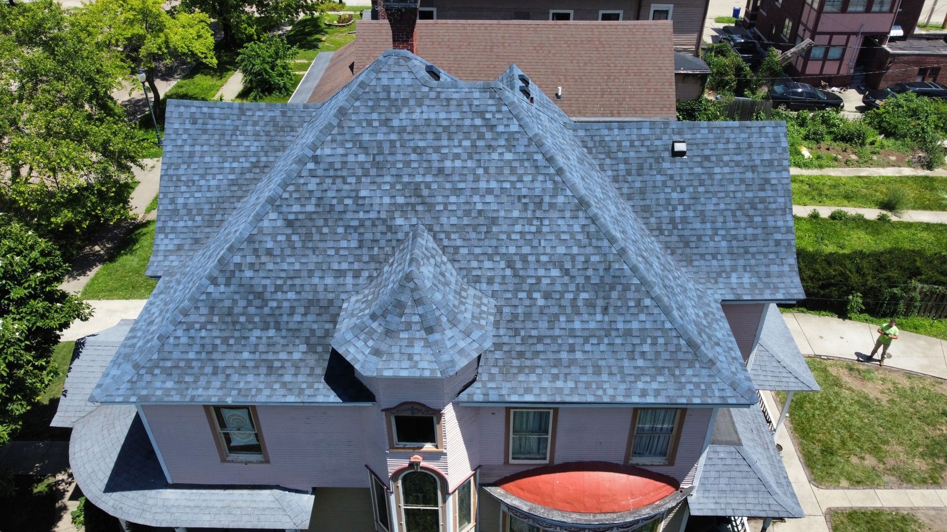 A high-angle aerial view of a historic home with a gray shingled roof, a turret, and a prominent red-roofed bay window.