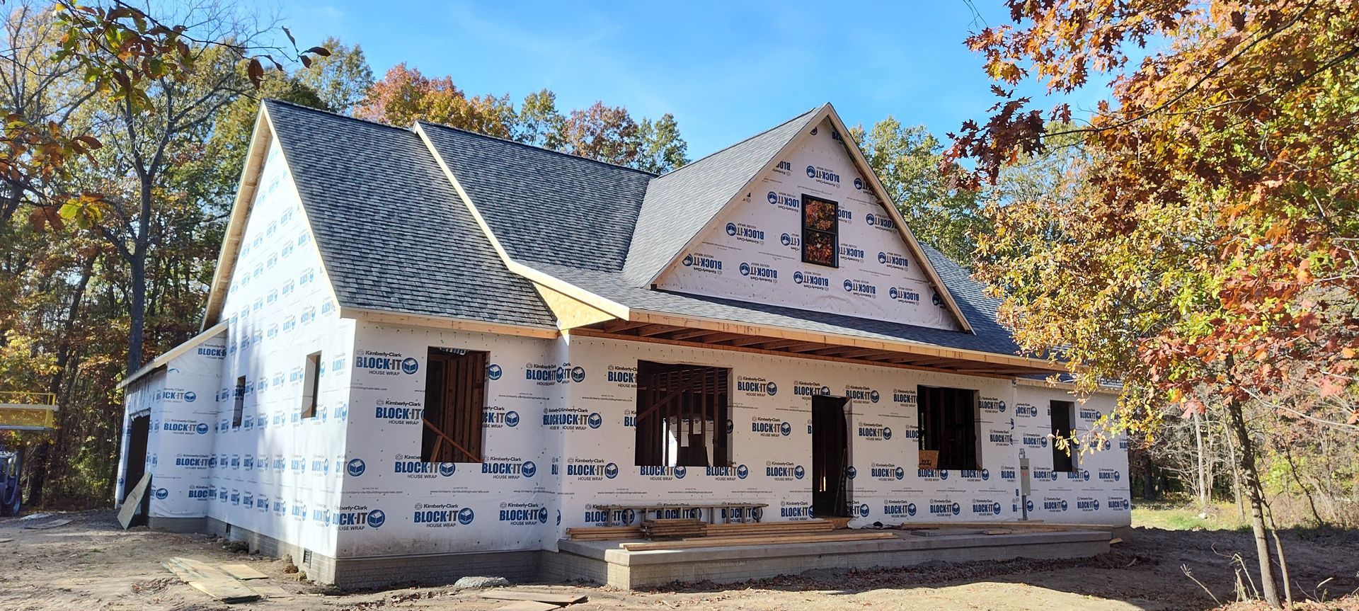 A new house under construction, wrapped in white weather-resistant barrier, with a shingled roof surrounded by trees.