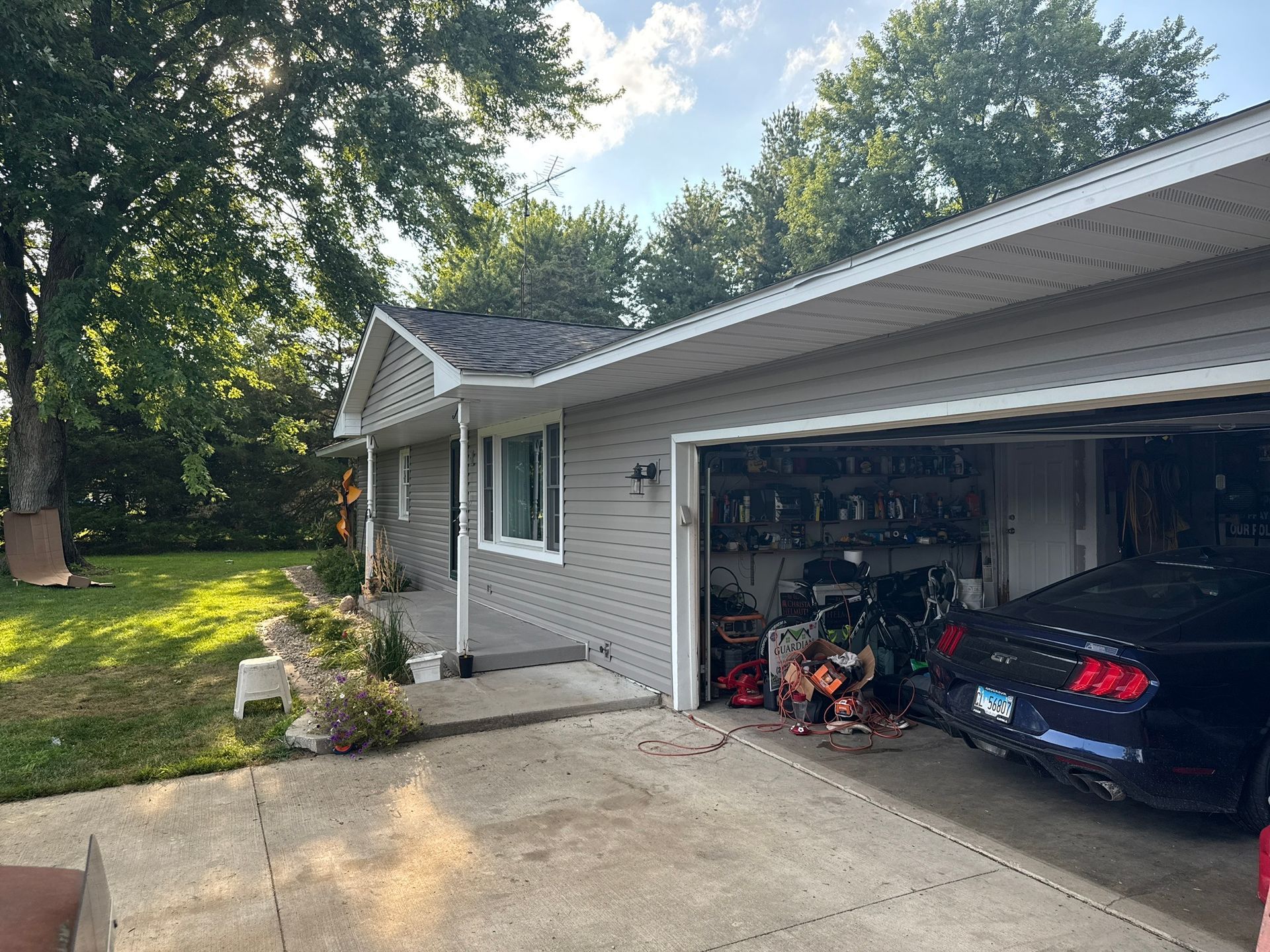A gray house with a wide driveway, an open two-car garage containing a dark sports car, and a green lawn with large trees.