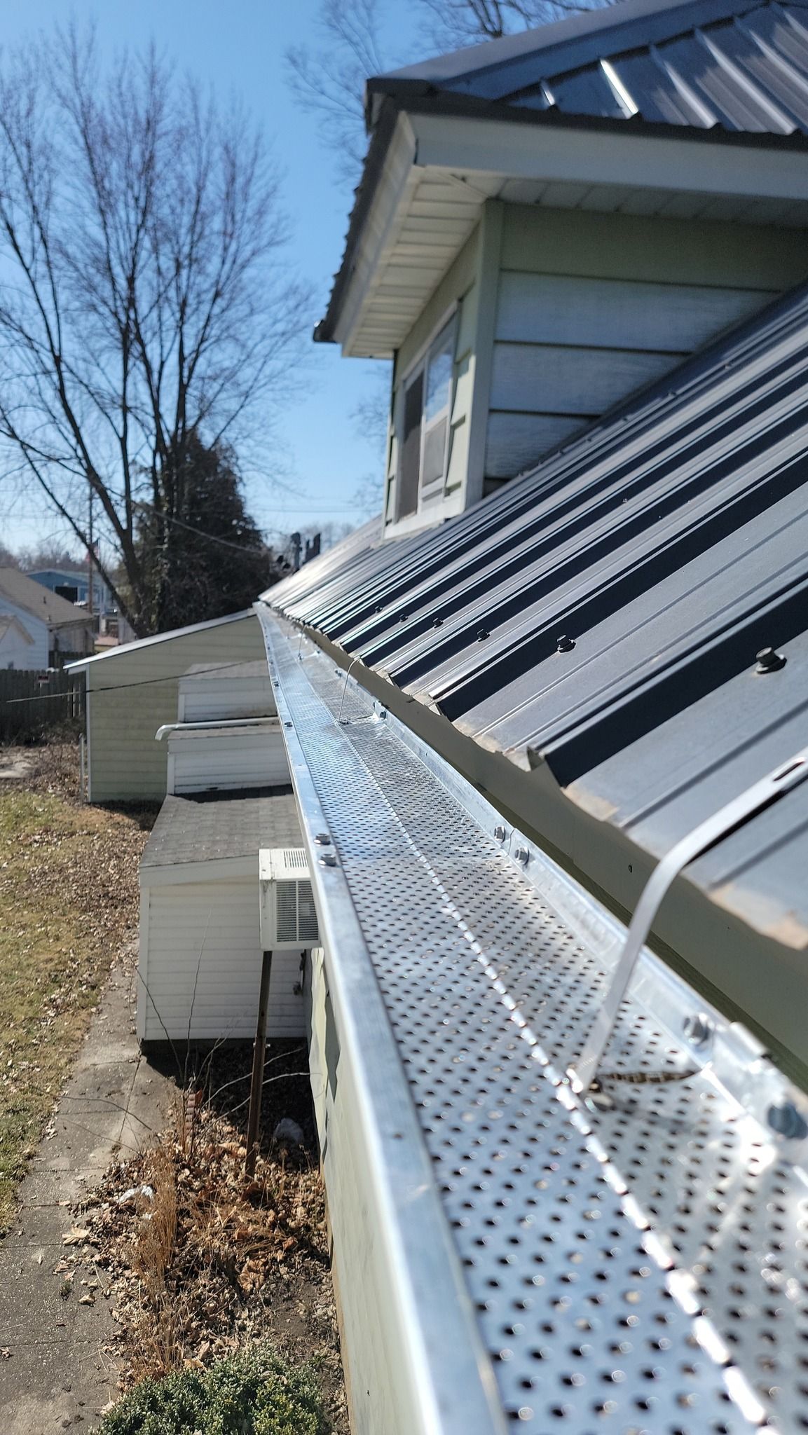 A metal gutter with a perforated screen cover runs along the edge of a house roof with dark metal shingles.