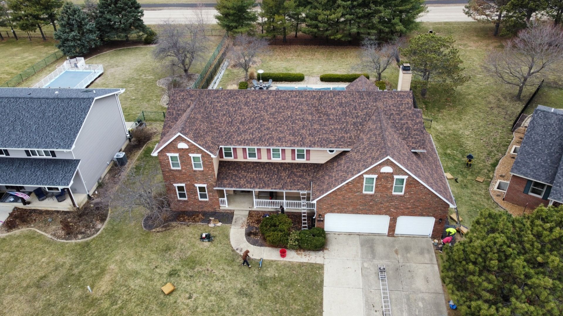 Aerial view of a two-story brick house with a dark shingled roof, surrounded by lawn, trees, and neighboring properties.
