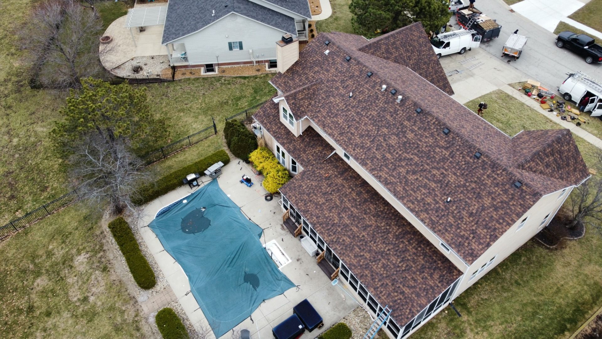 High-angle aerial view of a suburban house with a brown shingled roof, a covered swimming pool, and nearby parked trucks.