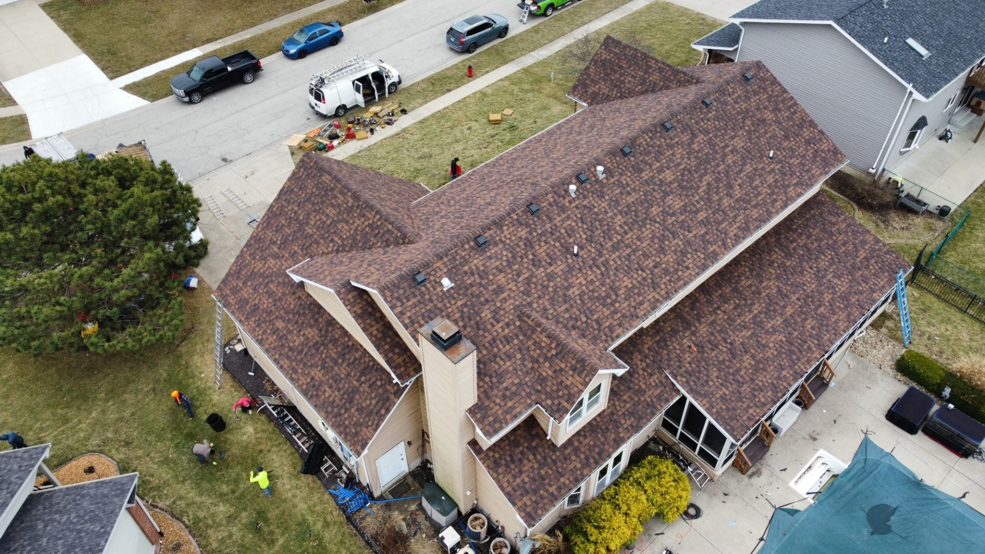 An aerial view of a multi-story house with a new brown shingled roof, surrounded by lawn, driveway, and parked vehicles.