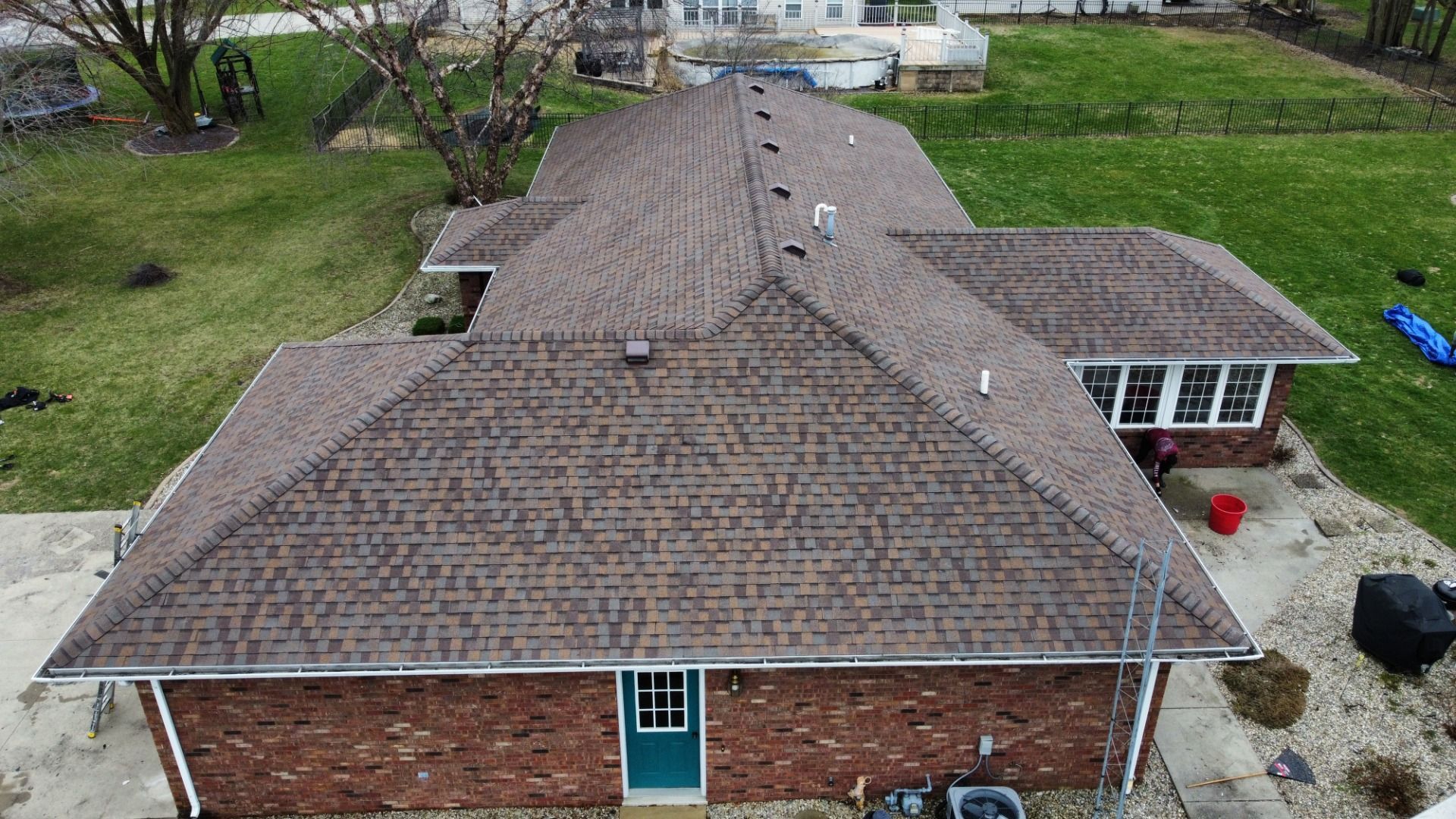 Aerial view of a residential brick house with a brown shingled hip roof, showing the backyard and surrounding lawn.
