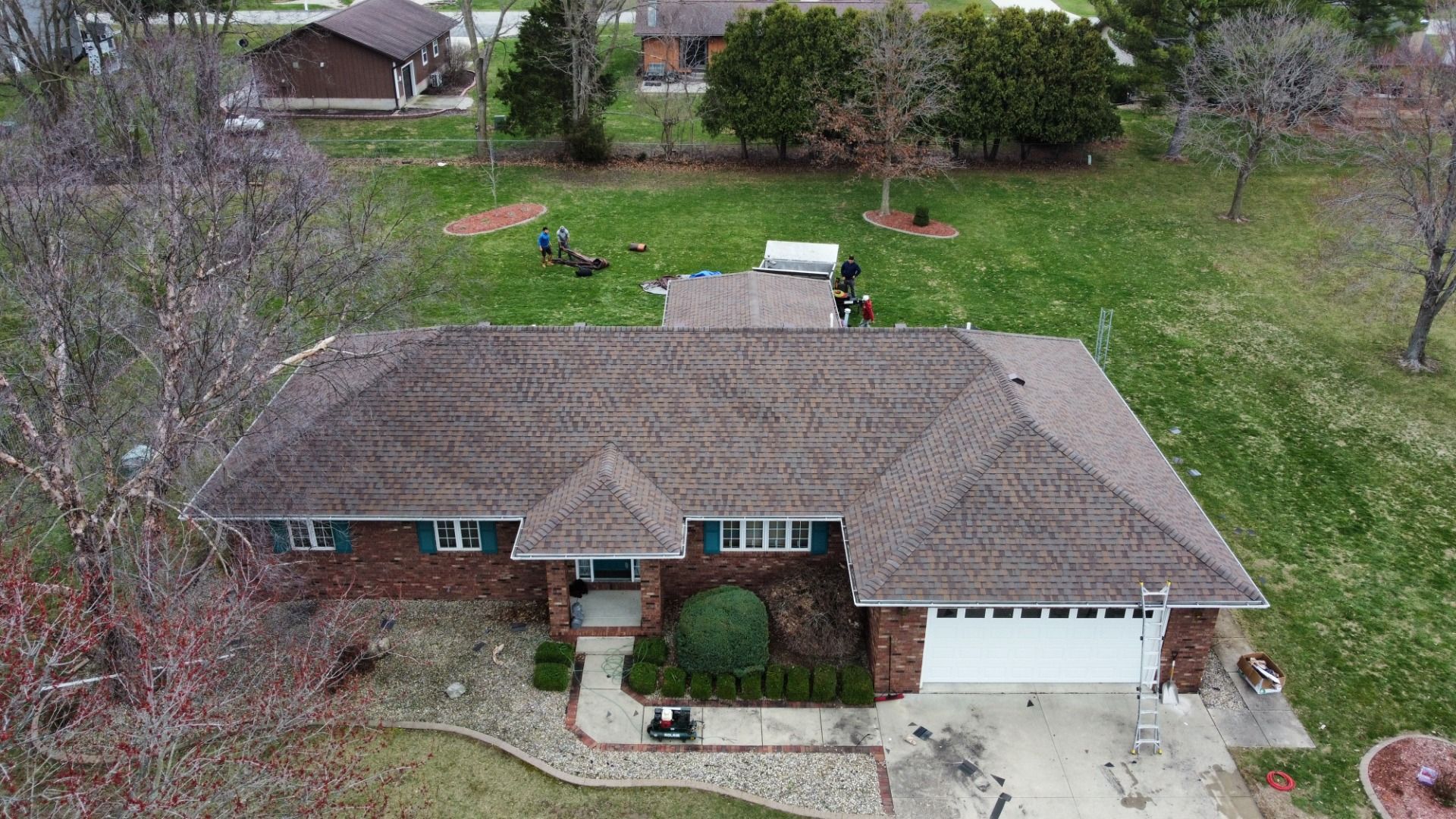 Aerial view of a brown brick ranch-style house with a gray shingled roof, surrounded by a lawn and trees.