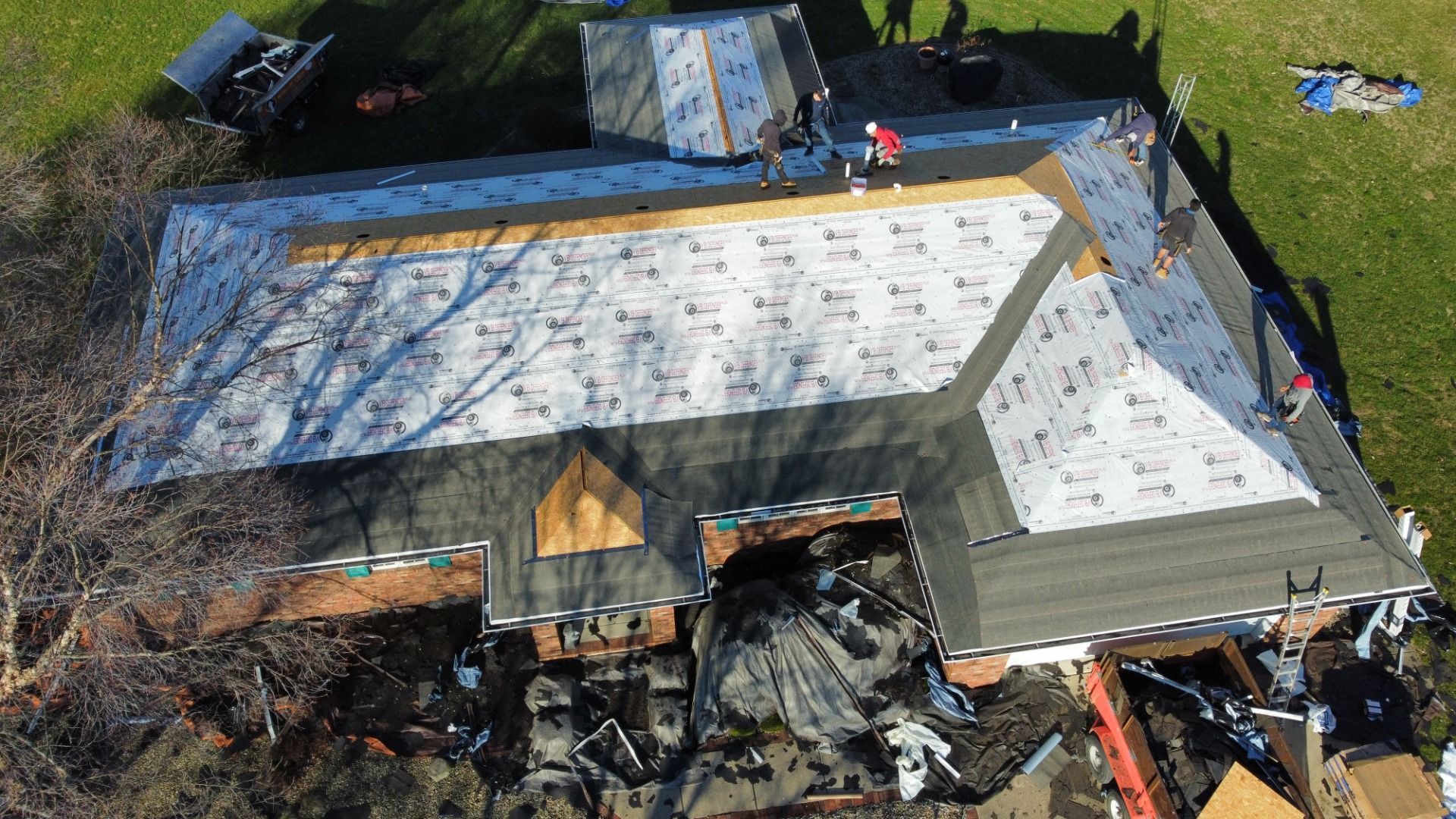 An aerial view shows workers installing new white underlayment on a house roof during a residential renovation project.