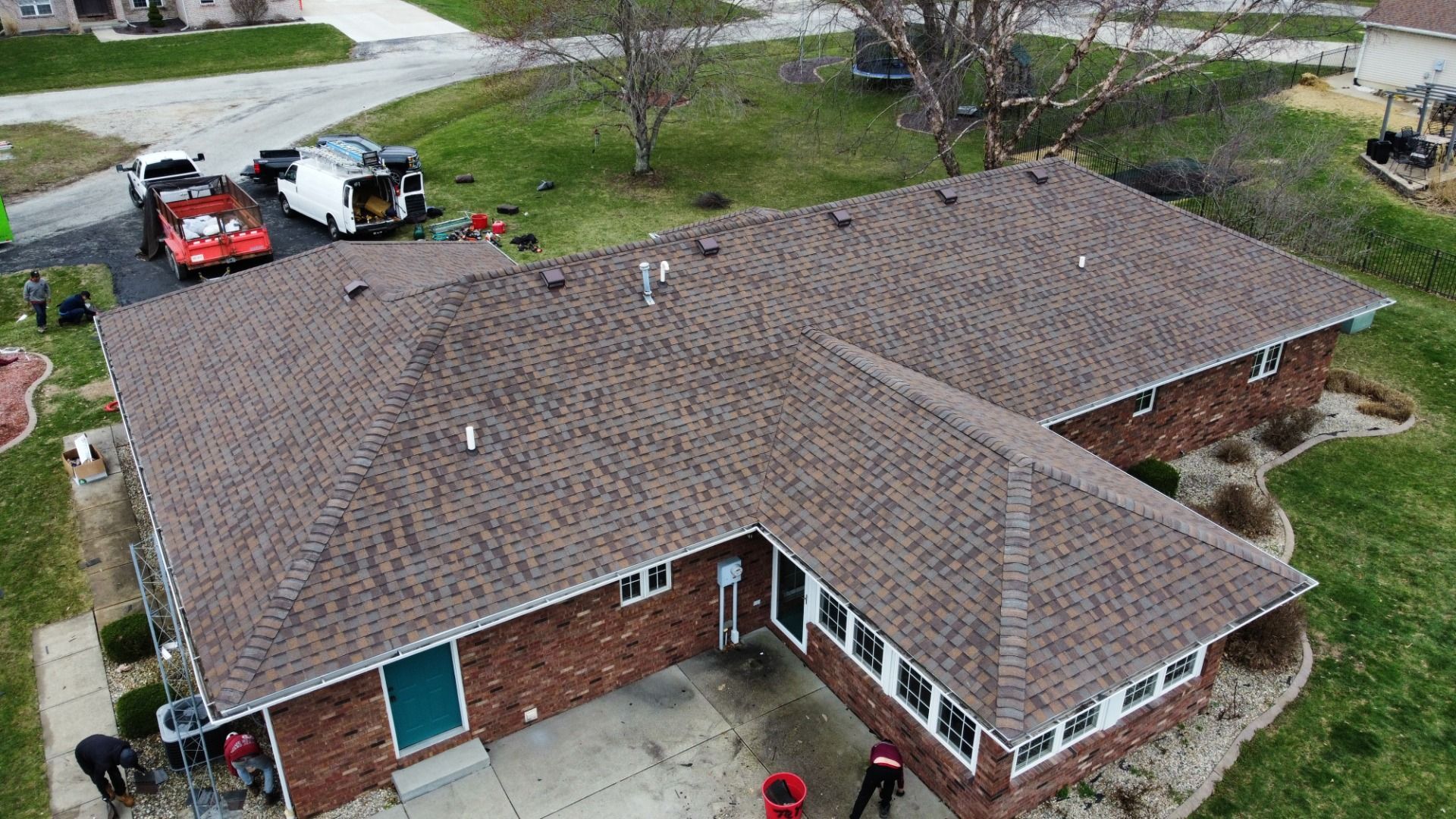 An aerial view of a brick house with a newly installed brown shingled roof, featuring work crews on the ground.