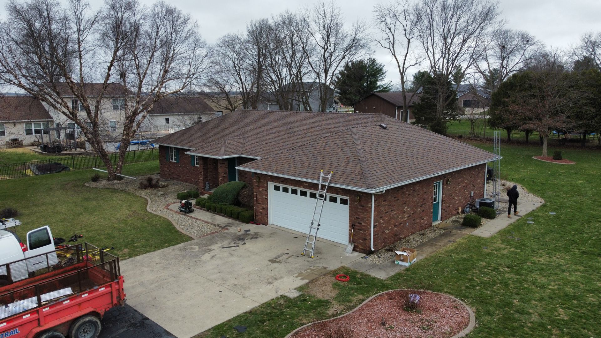 An elevated view of a brick house with a new shingled roof, a ladder against the garage, and a truck parked in the yard.