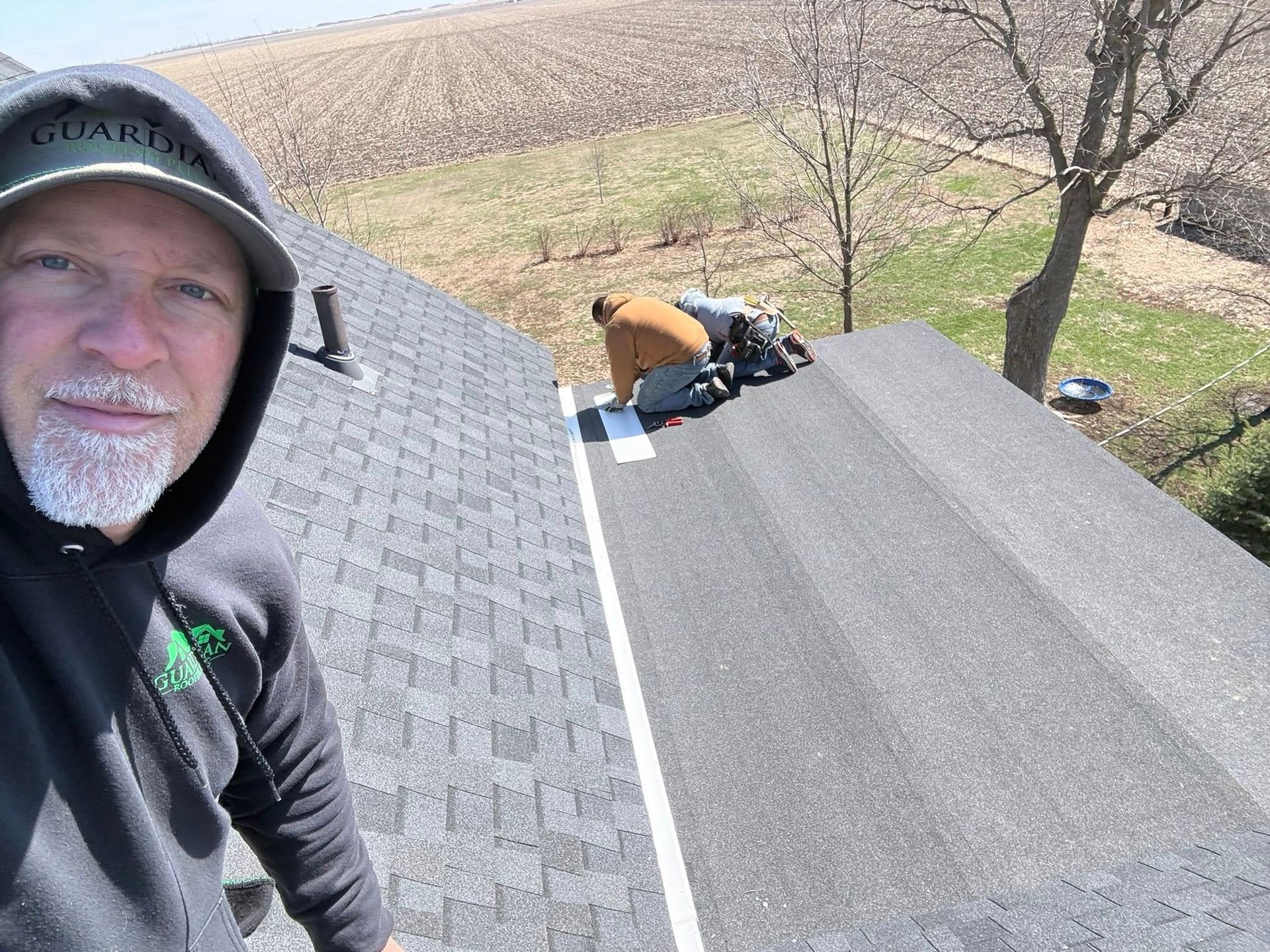 A person in a hooded sweatshirt selfies on a roof while another person works on shingles in the background.