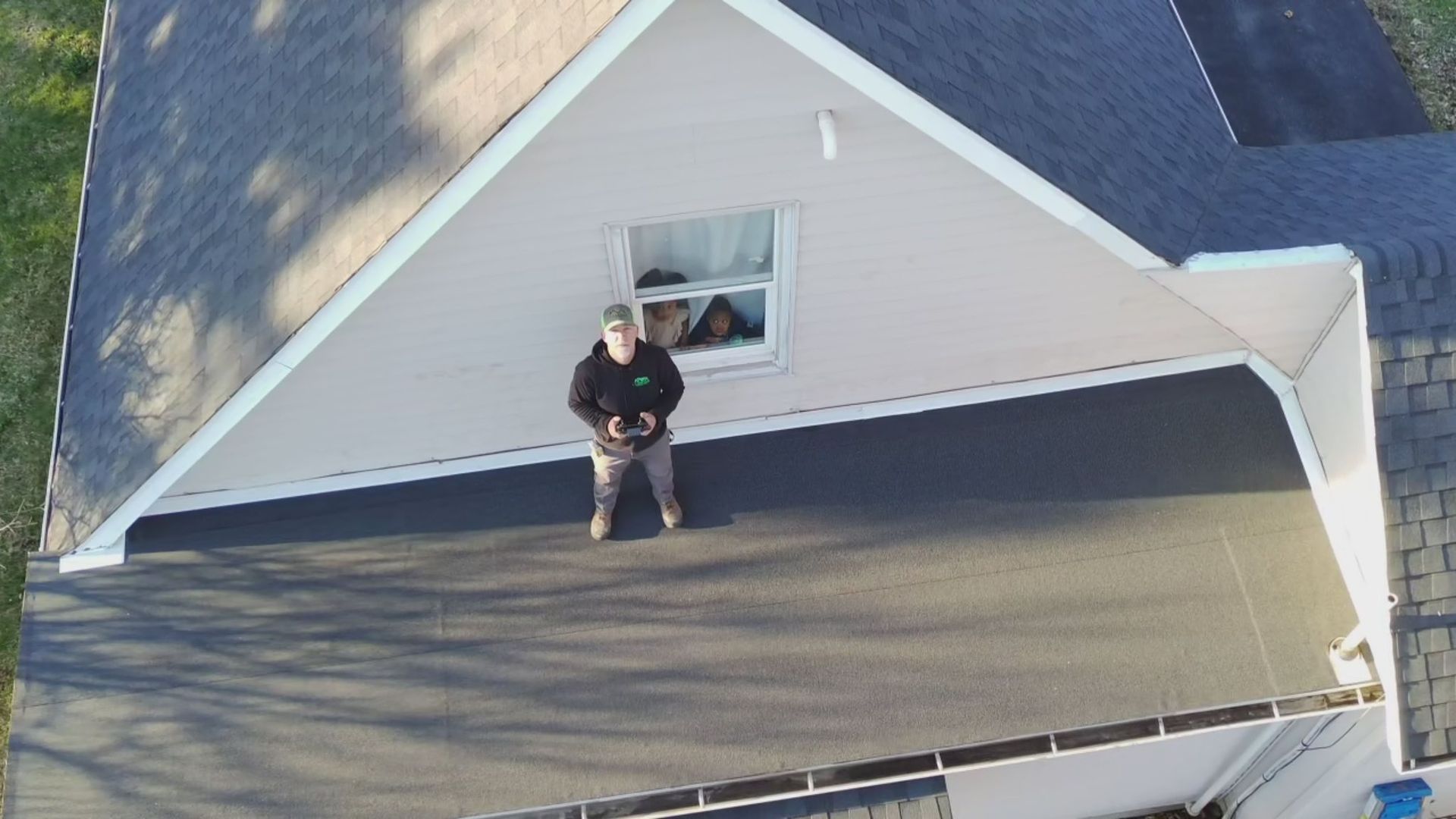 A person holding a drone controller stands on a flat, dark roof next to a house wall with a window.