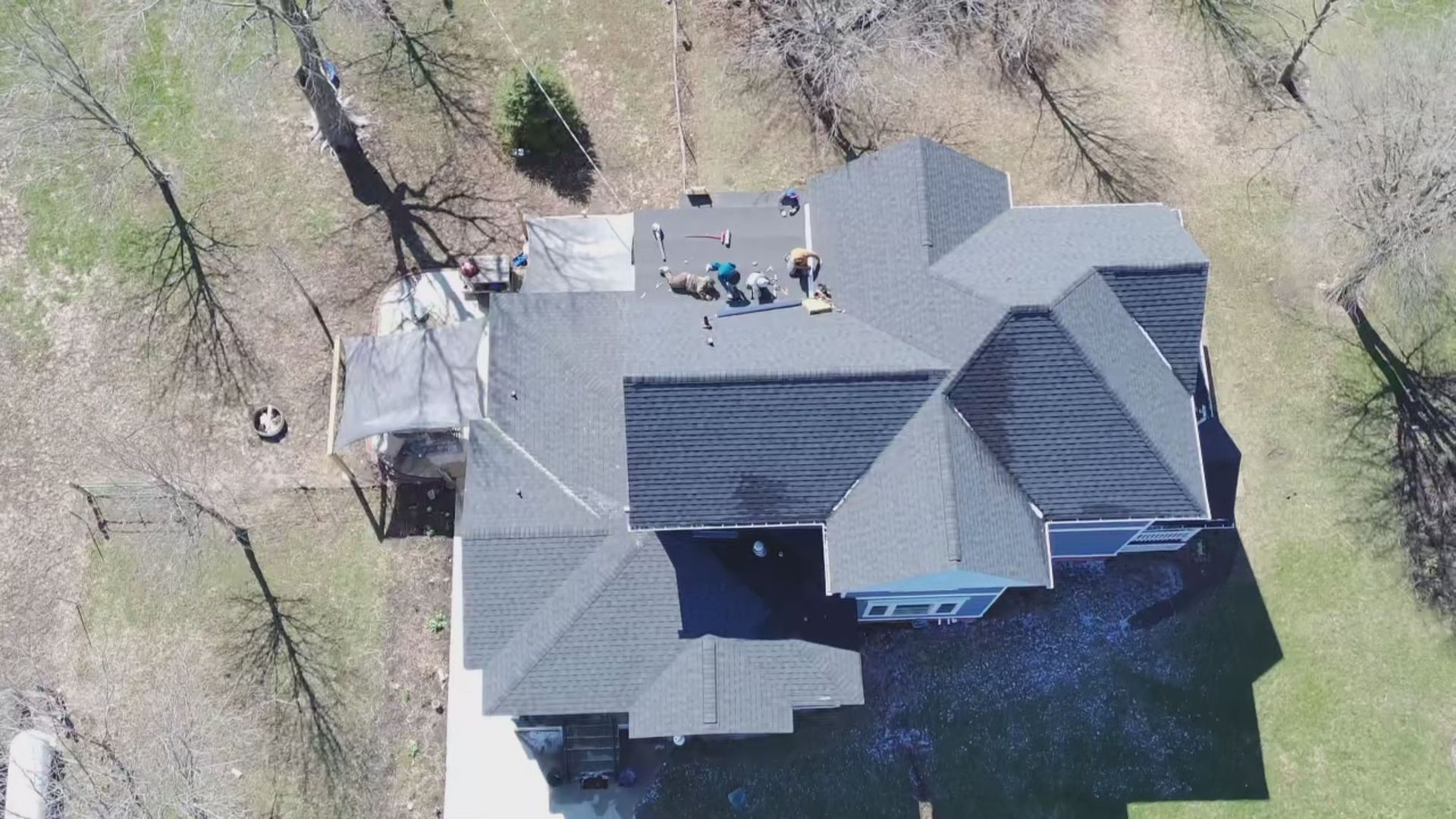 An aerial view of a gray shingled residential roof with workers performing repairs on a sunny day.