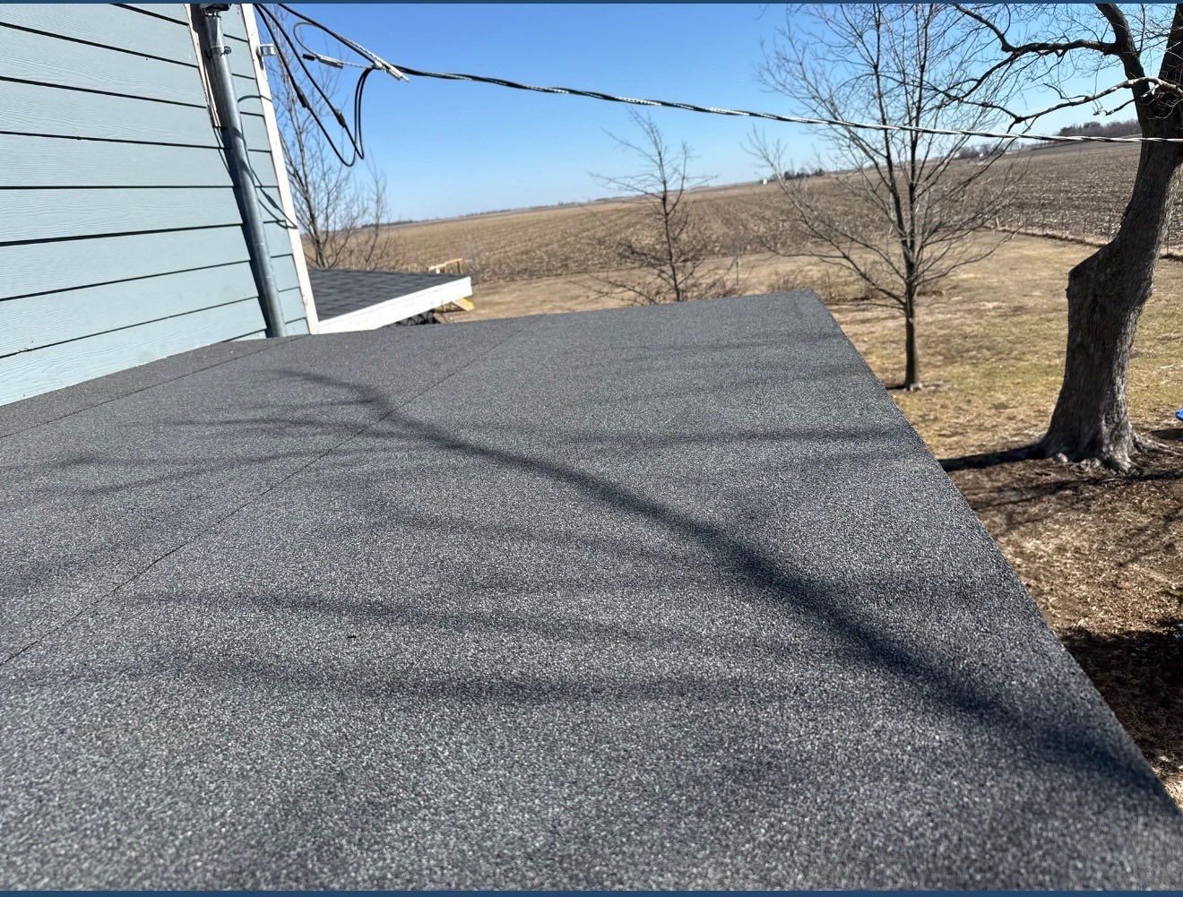 A high-angle view of a flat, gray, granular-textured roof surface extending from the side of a light-blue house.