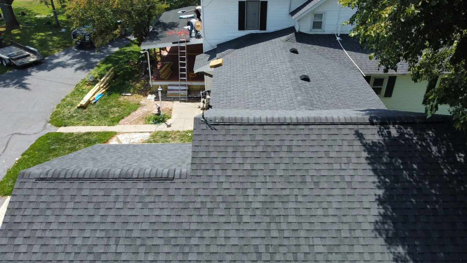 An aerial view of a dark grey shingled roof on a house, with a ladder leaning against a side porch and nearby grass.