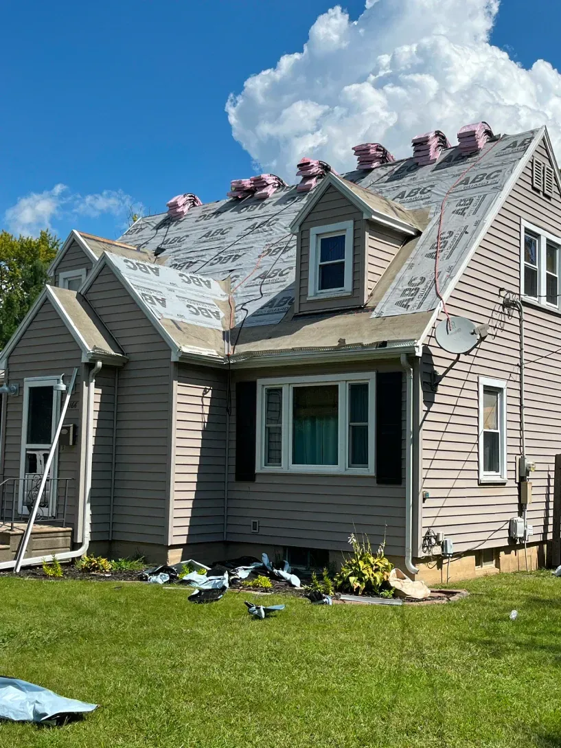 A single-story house with beige siding undergoing roof repairs, with visible roofing underlayment and debris on the lawn.