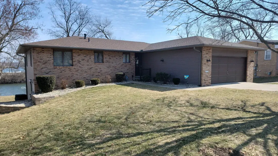 A single-story, L-shaped house with a tan brick exterior, brown siding, a two-car garage, and a large lawn.