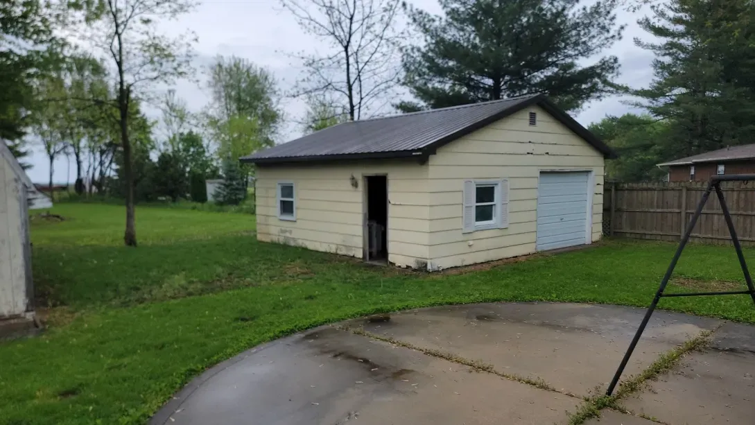 A light yellow detached garage with a dark roof, side door, and single white garage door sits in a grassy backyard.