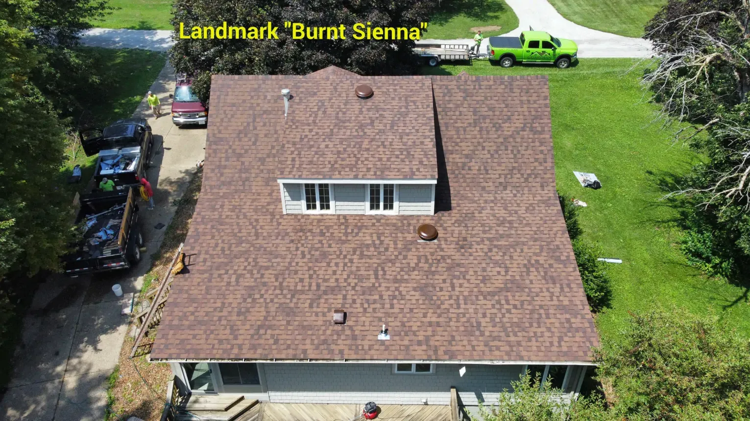 An aerial view of a house with a new Burnt Sienna shingle roof, with construction vehicles parked in the driveway.