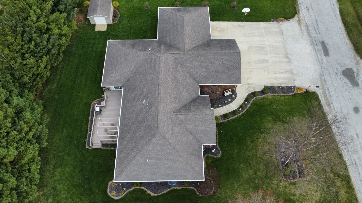 A top-down aerial view of a single-story house with a gray shingled roof, surrounding lawn, driveway, and small shed.