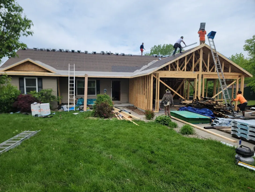 Roofers work on the shingled roof of a house with a new wooden frame structure attached to the side.