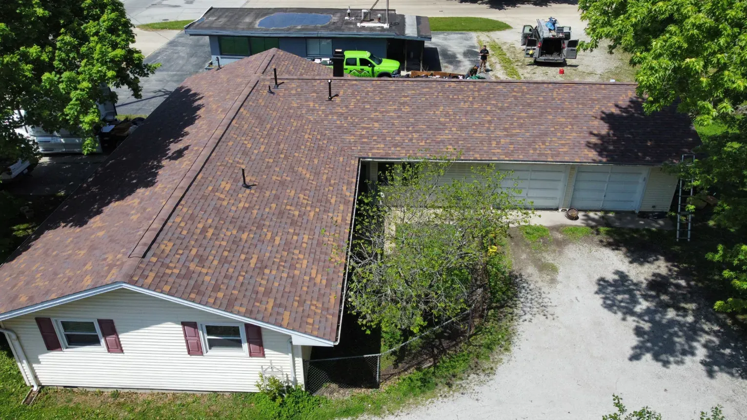 Aerial view of a residential home with a brown shingled roof, white siding, and a gravel driveway on a sunny day.