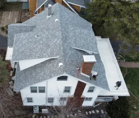 Aerial view of workers installing gray shingles on a multi-pitched roof with a brick chimney and a flat white roof section.