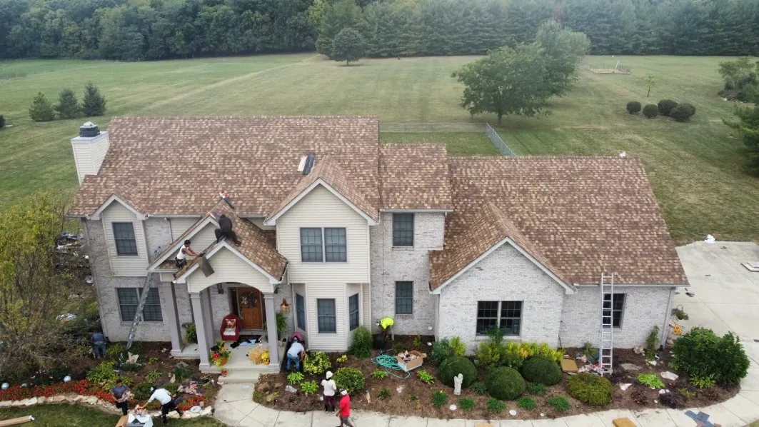 Aerial view of a two-story stone house with a brown shingled roof, surrounded by landscaping, with workers near the base.