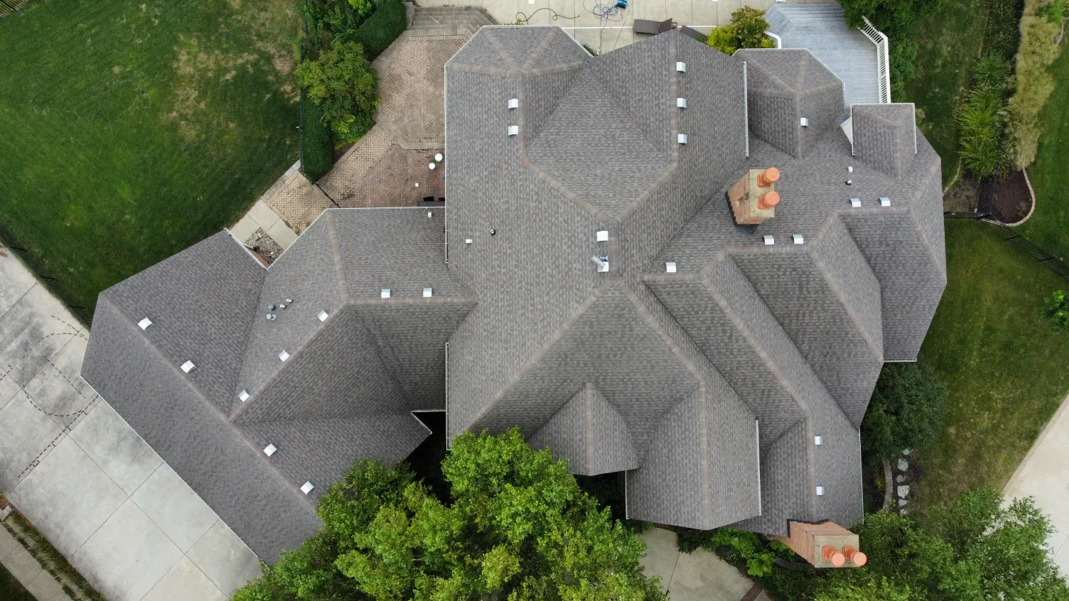 An aerial top-down view of a complex, multi-gabled gray shingled roof on a residential home surrounded by lawn and trees.