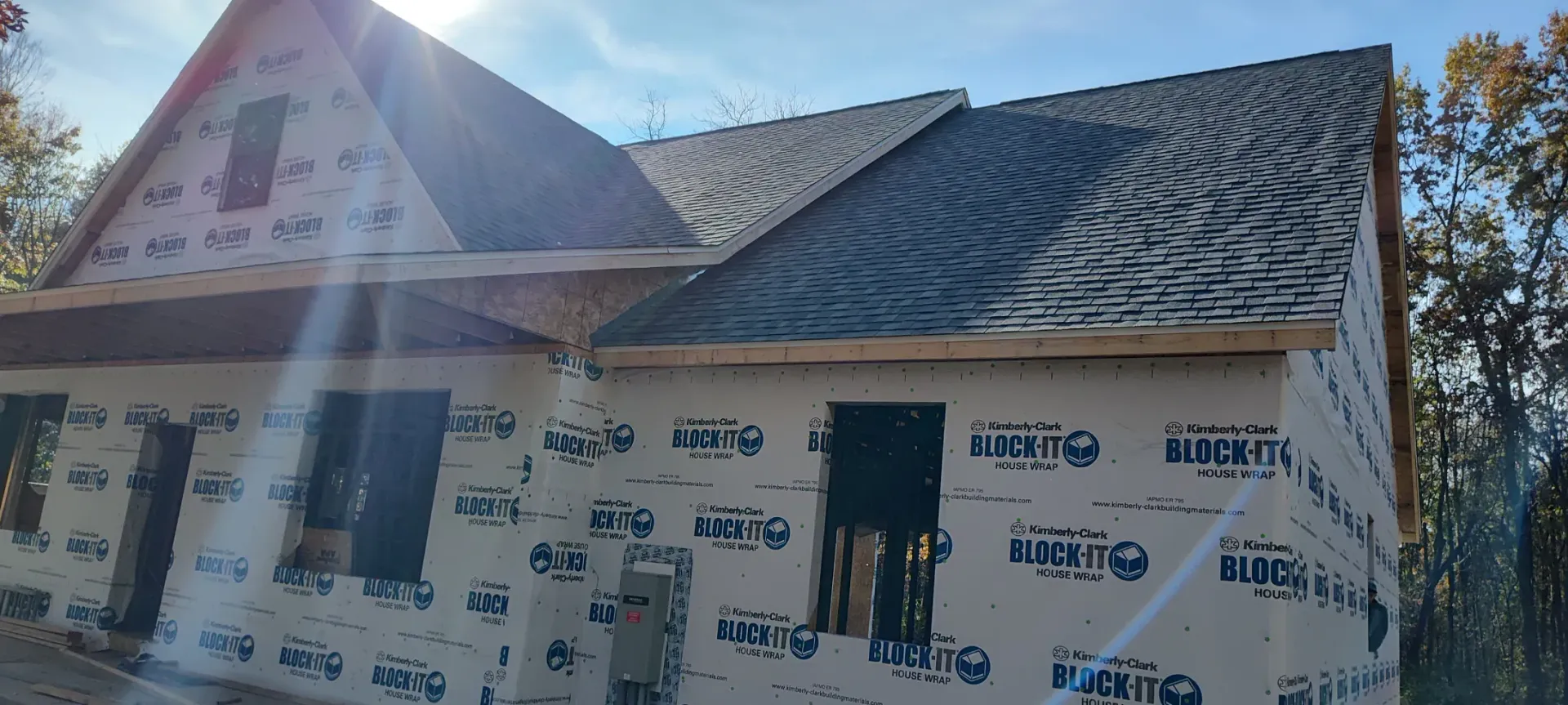 A partially constructed house exterior wrapped in white weather barrier with shingled roof sections under a bright sun.