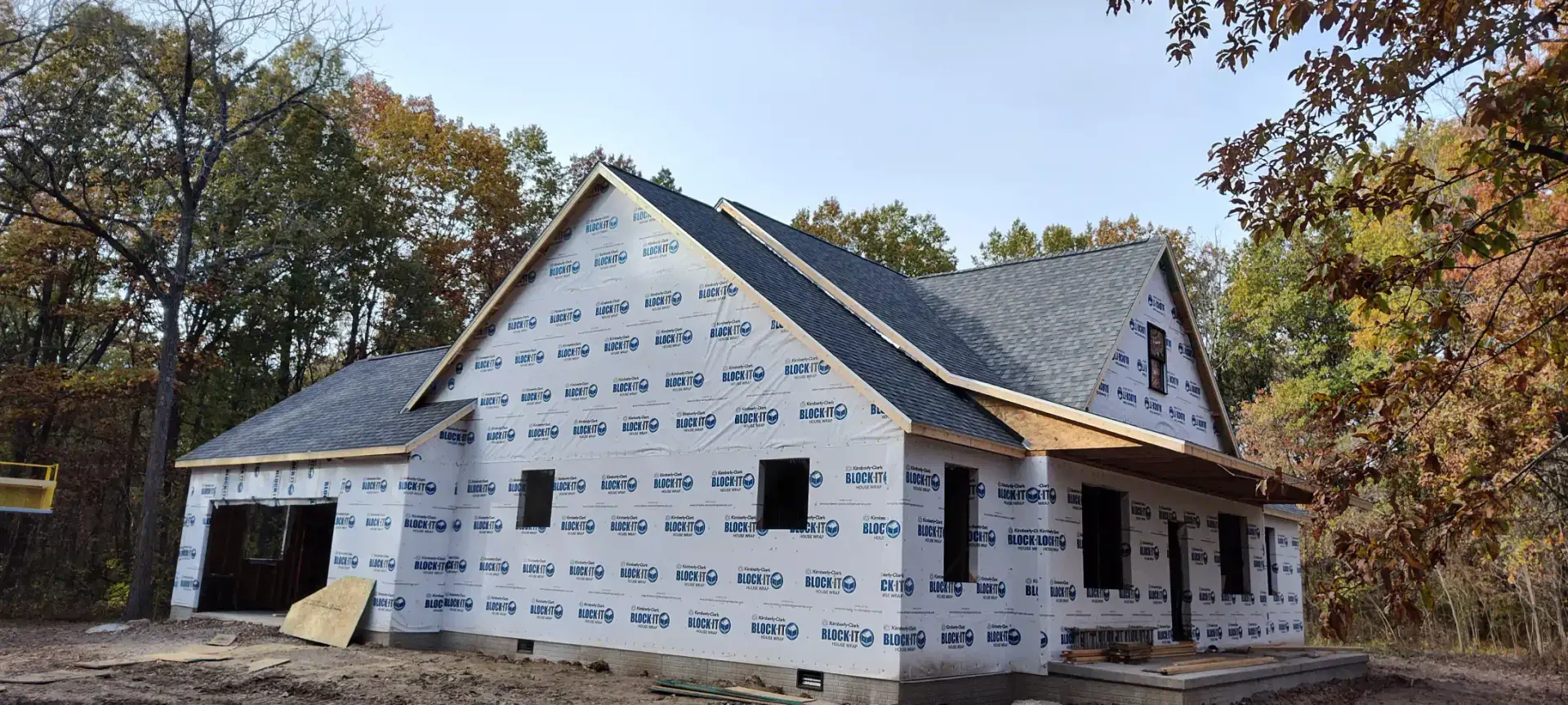 A new house under construction, covered in white protective wrap, with a shingled roof surrounded by autumn trees.