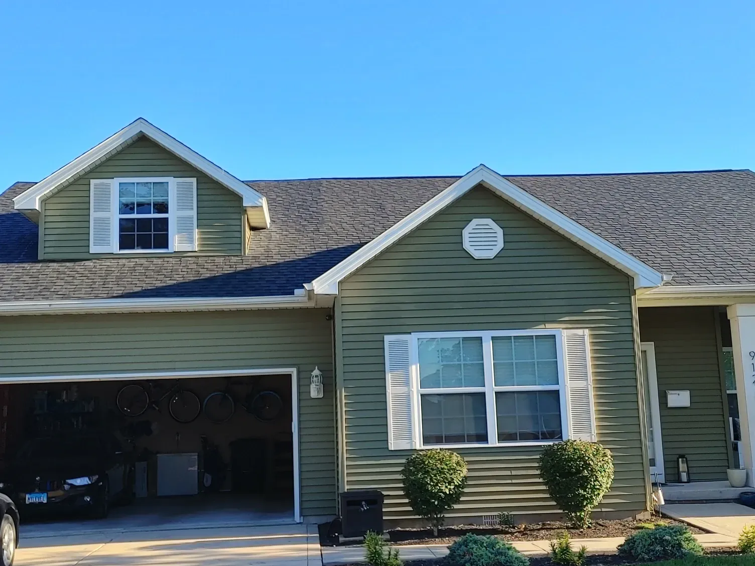 A two-story olive green house with a garage, white trim, and a shingled roof under a clear blue sky.