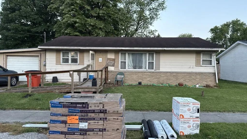 A single-story beige house with a wheelchair ramp and roofing supplies stacked on the lawn in front.
