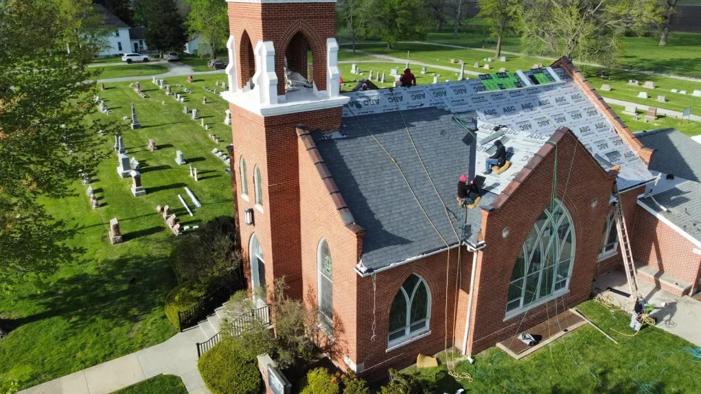 Aerial view of a red-brick church with workers repairing the roof, located next to a graveyard on a sunny day.