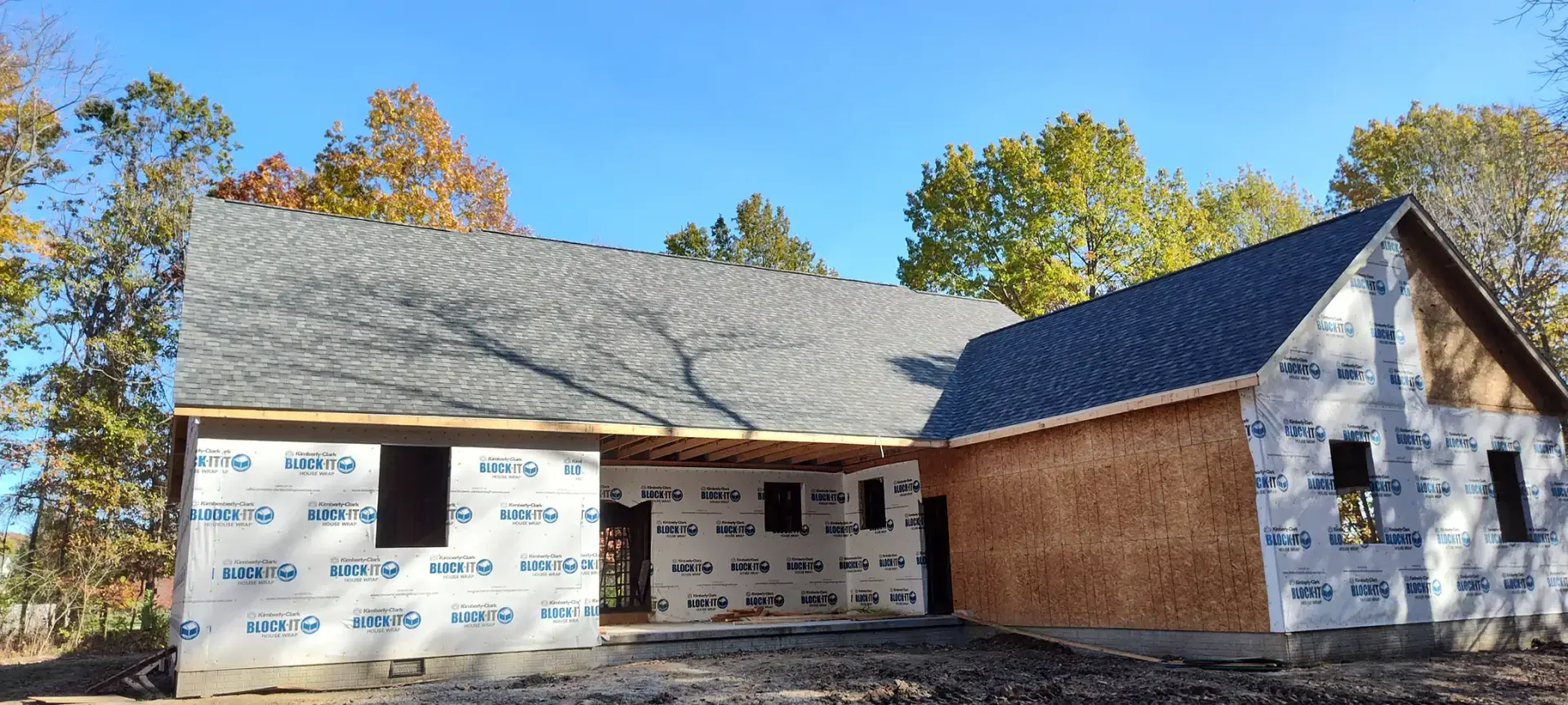 A partially constructed house frame under a clear blue sky, featuring unfinished walls, wooden panels, and a shingled roof.