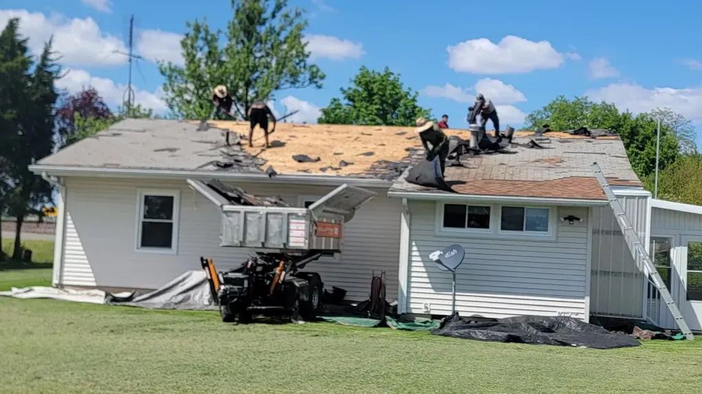 Workers in dark clothing removing old shingles from a house roof into a large metal dumpster parked in the yard.