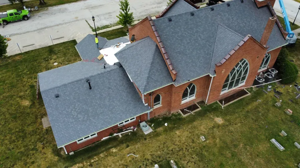 An aerial view of a brick church undergoing roof repair, showing gray shingles and workers near a cemetery.