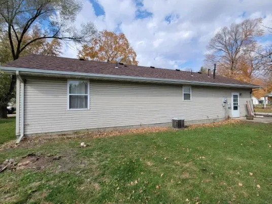 Side view of a single-story home with light beige vinyl siding, a brown shingled roof, and green lawn.