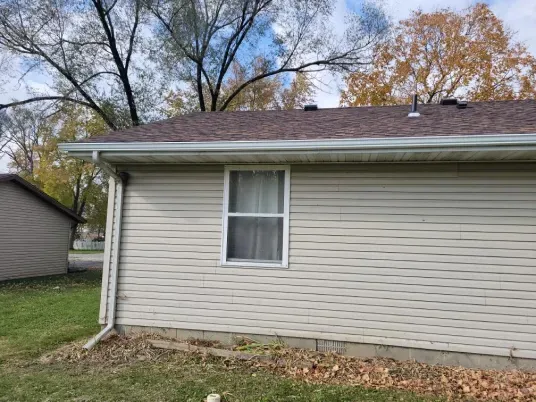 Exterior view of a beige vinyl-sided house with a shingled roof, a single window, and a white gutter and downspout.