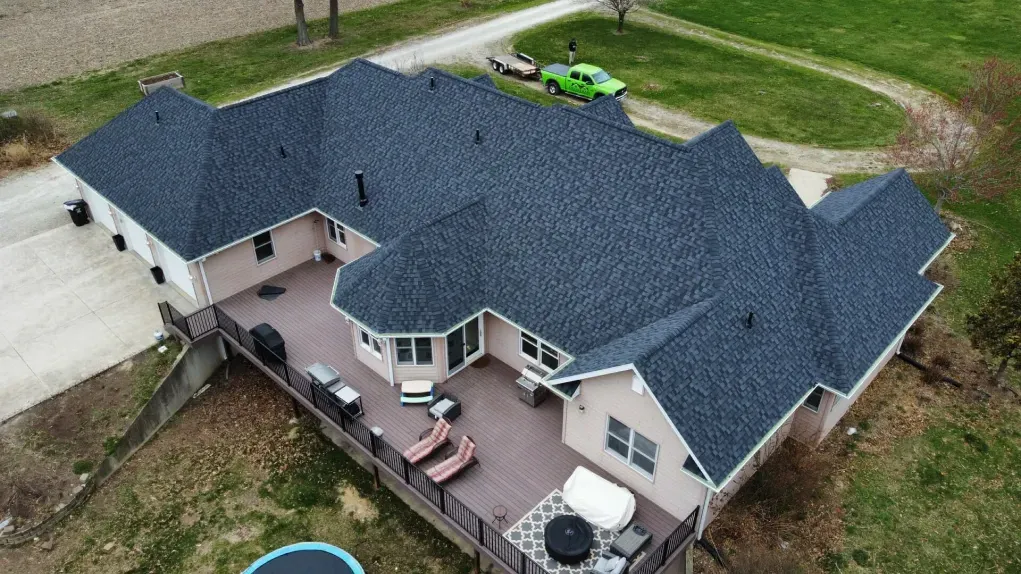An aerial view of a light-colored ranch-style home with a dark gray shingled roof and a spacious deck in a rural setting.