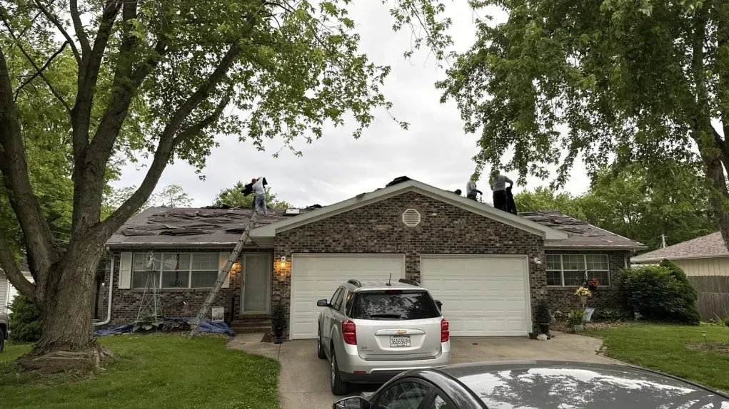 Workers on the roof of a stone-faced single-story home with a two-car garage and two vehicles parked in the driveway.