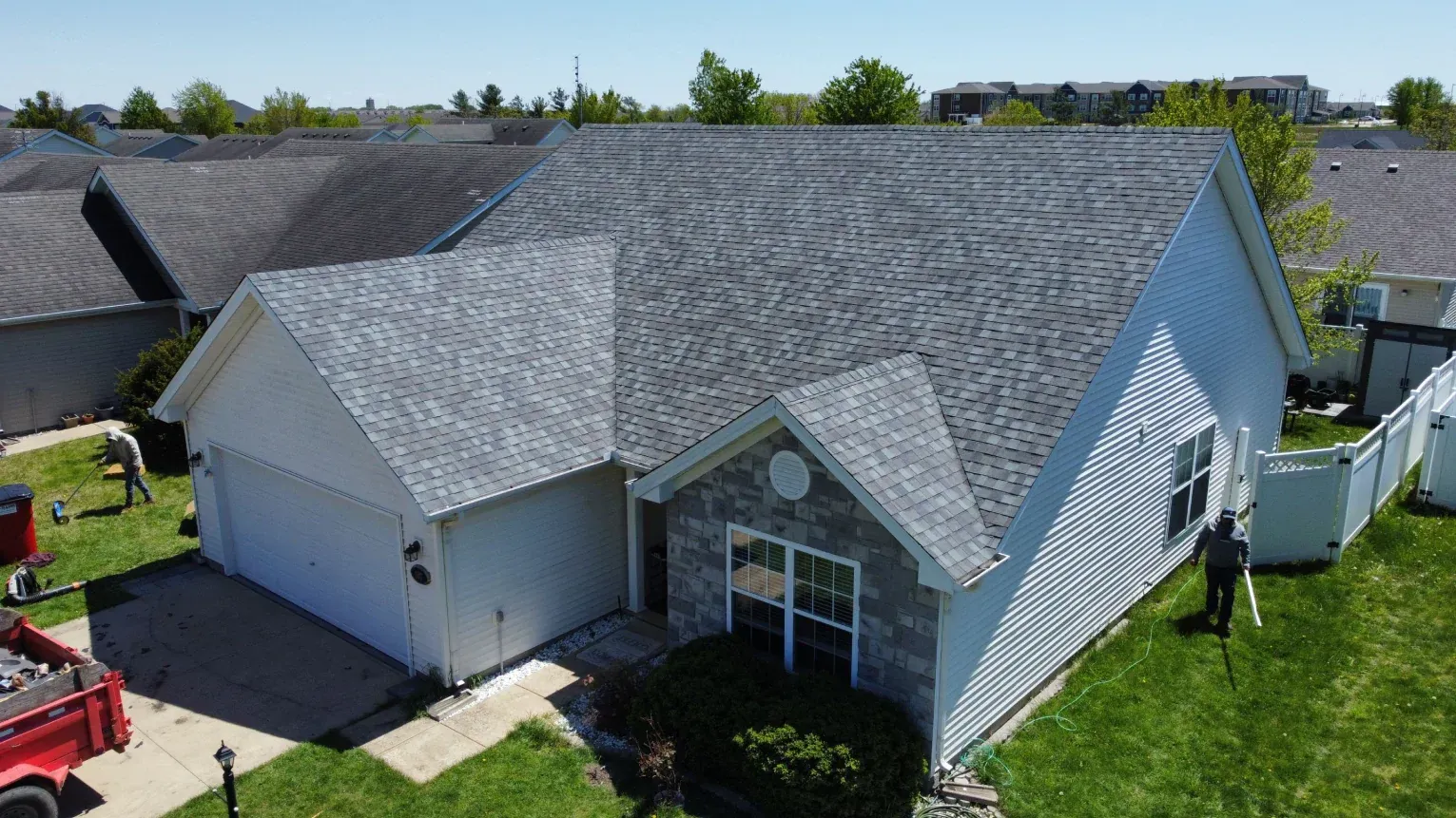 An aerial view of a single-story suburban house with a gray shingled roof, white siding, and a stone front, during roofing.