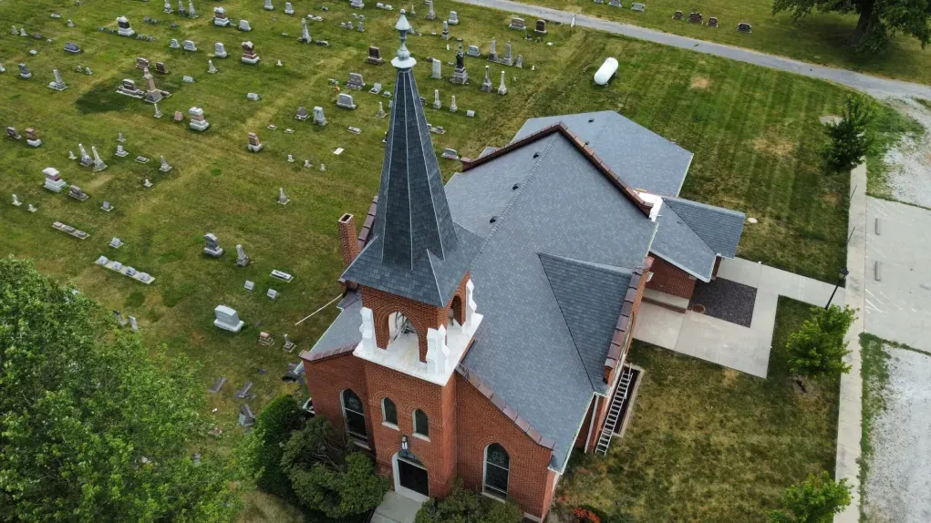 An aerial view of a red brick church with a steeple, situated beside a cemetery filled with gravestones.