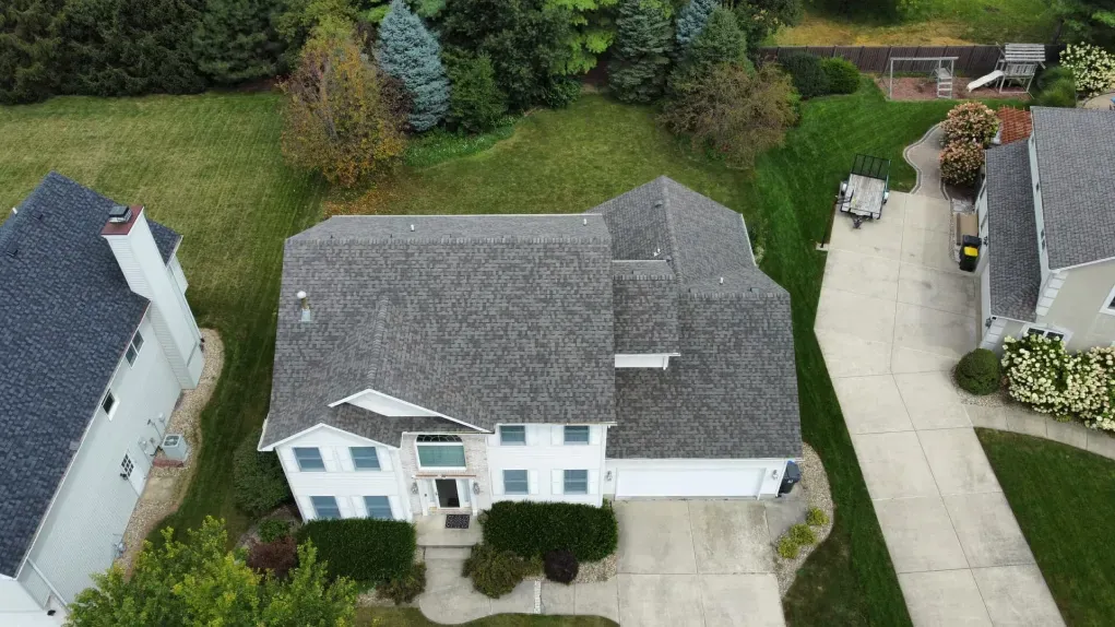 An aerial, top-down view of a white two-story suburban house with a gray shingled roof, lawn, and concrete driveway.