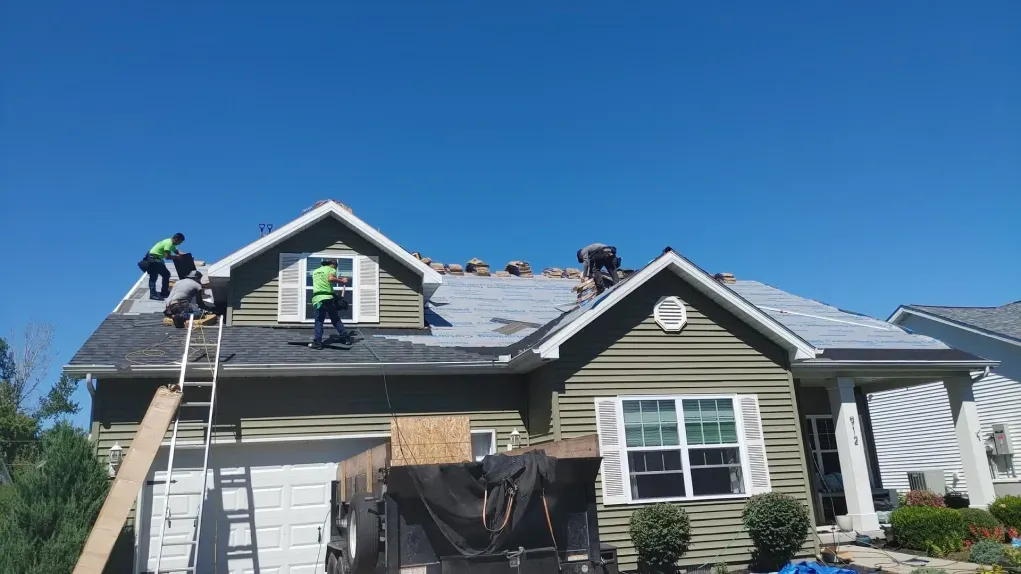 Three workers in high-visibility vests repair a green house's shingled roof under a clear blue sky.