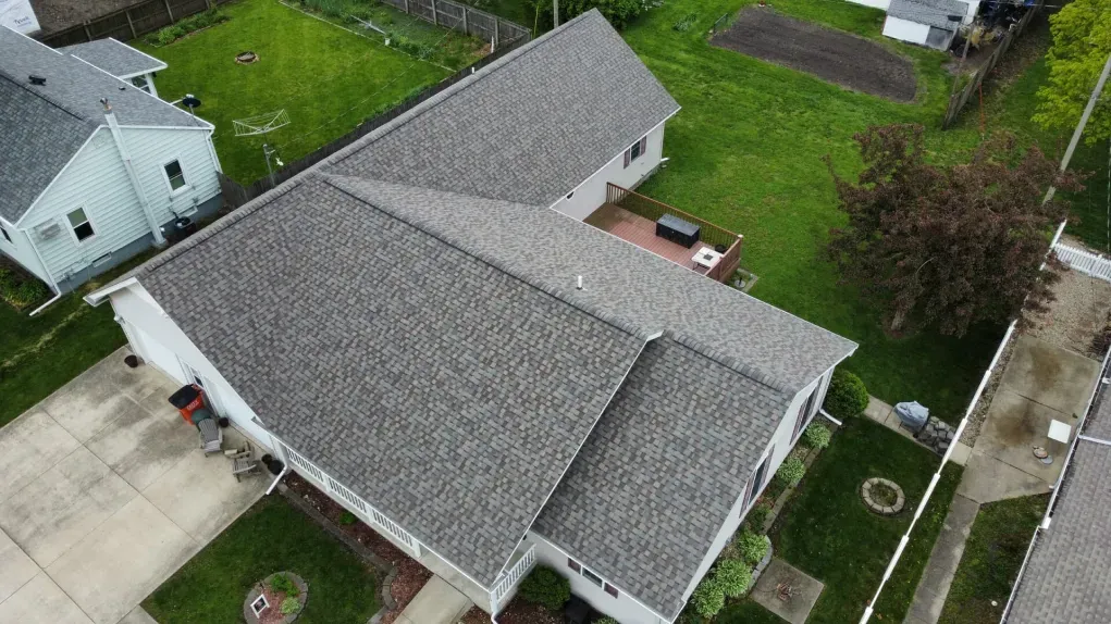 High-angle aerial view of a gray-shingled L-shaped residential roof on a suburban property with a yard and driveway.