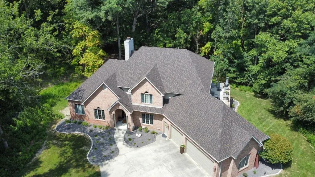 An aerial view of a tan brick house with a dark gray shingled roof, surrounded by green trees and a paved driveway.