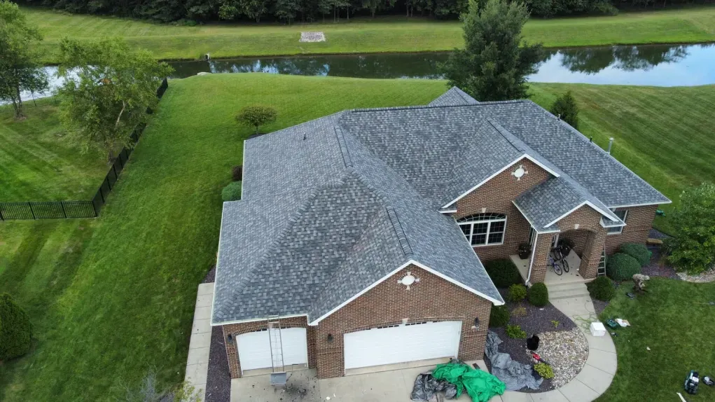An aerial view of a brick house with a dark shingled roof, a multi-car garage, and a large lawn bordered by a water feature.
