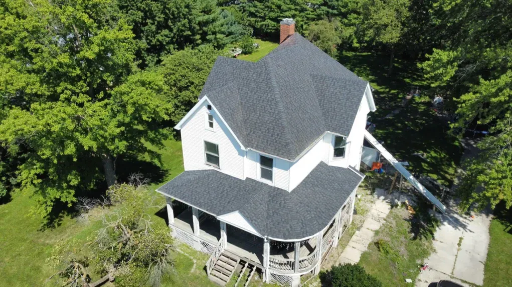 An aerial view of a white two-story house with a grey shingled roof, surrounded by green trees and a paved driveway.