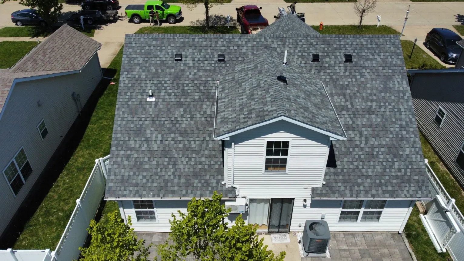 An aerial view of a suburban house with a gray shingled roof, white siding, and a small backyard with trees and a patio.