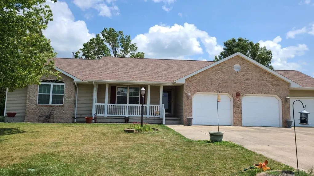A single-story tan brick house with a white front porch and a two-car garage under a brown shingled roof.