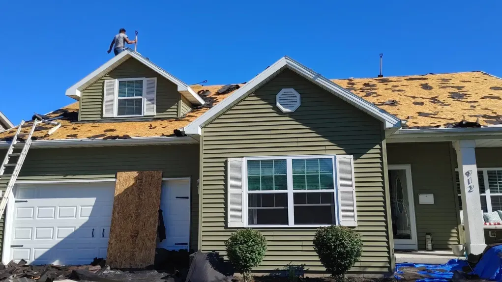 A person works on the roof of a green house with exposed plywood under a clear blue sky.